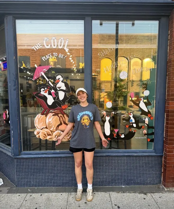 Storefront window painted with two cartoon penguins lounging with tropical drinks under hand-lettered text reading "The Cool Place to Be"