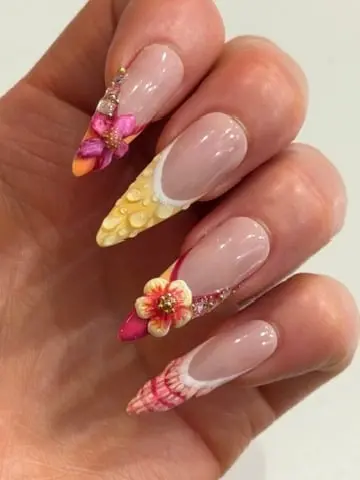 Close-up of long stiletto acrylic nails with a nude French tip, accented by hand-sculpted 3D pink and orange flowers and a textured yellow dewdrop design